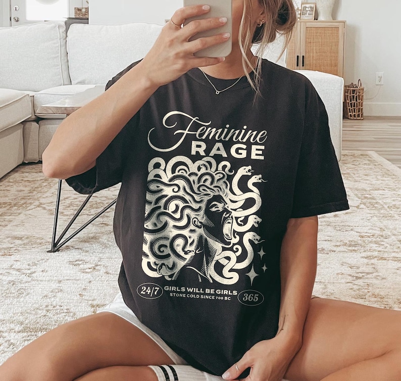 May include: Black t-shirt with a white graphic of a woman with snakes for hair. The text "Feminine Rage" is above the graphic and "Girls will be girls. Stone cold since 700 BC" is below the graphic.