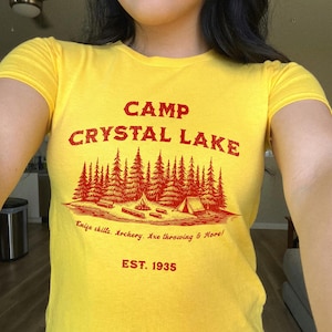 May include: Yellow t-shirt with red text that reads "Camp Crystal Lake" and "Knife skills. Archery. Axe throwing & More! EST. 1935".