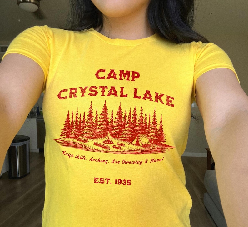 May include: Yellow t-shirt with red text that reads "Camp Crystal Lake" and "Knife skills. Archery. Axe throwing & More! EST. 1935".
