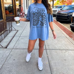 May include: A light blue t-shirt with a graphic of a woman's face with snakes around her head. The text "Feminine Rage" is printed on the shirt. The text "24/7" and "365" are printed on the bottom of the shirt.