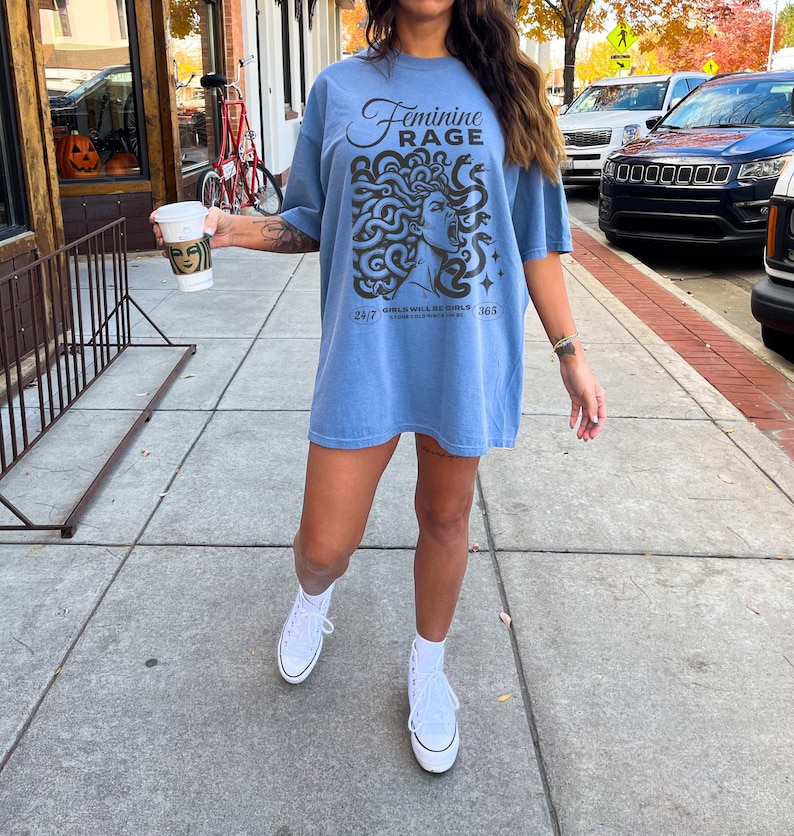 May include: A light blue t-shirt with a graphic of a woman's face with snakes around her head. The text "Feminine Rage" is printed on the shirt. The text "24/7" and "365" are printed on the bottom of the shirt.