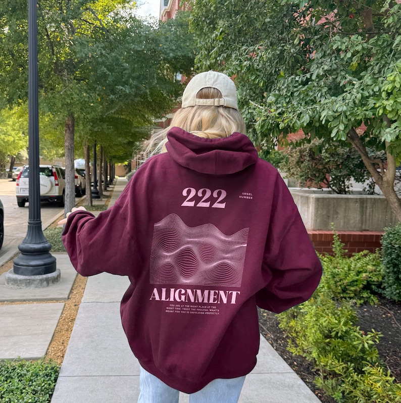 May include: A maroon hoodie with a graphic design on the back. The graphic features a wavy line pattern and the text "222 Angel Number" and "Alignment".