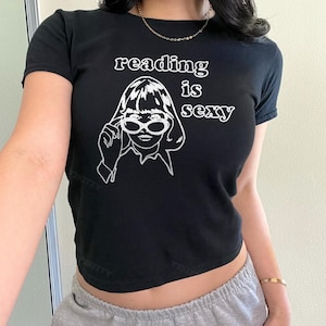 May include: Black crop top with the text "reading is sexy" and a white graphic of a woman wearing sunglasses. The shirt has short sleeves and a round neckline. The overall style is casual and trendy.