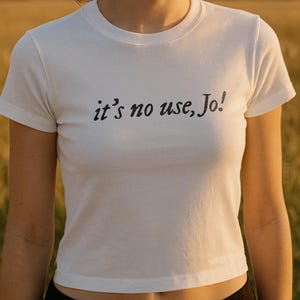 May include: White crew neck t-shirt with the phrase "it's no use, Jo!" printed in black. The shirt is short-sleeved and appears to be made of a soft material. The model is wearing black pants.