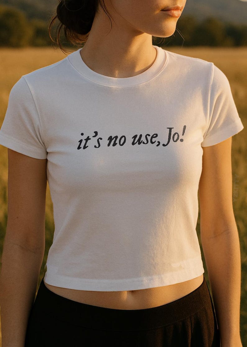 May include: White crew neck t-shirt with the phrase "it's no use, Jo!" printed in black. The shirt is short-sleeved and appears to be made of a soft material. The model is wearing black pants.