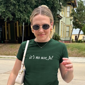 May include: A dark green t-shirt with the text "it's no use, Jo!" in white. The person is wearing gold-rimmed sunglasses, gold hoop earrings, and a delicate necklace. A white purse with a gold chain is visible.