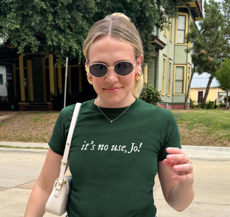 May include: A dark green t-shirt with the text "it's no use, Jo!" in white. The person is wearing gold-rimmed sunglasses, gold hoop earrings, and a delicate necklace. A white purse with a gold chain is visible.