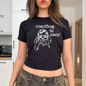 May include: Black t-shirt with the white text "reading is sexy" above a line drawing of a woman wearing sunglasses. The woman is touching her glasses. The shirt is short-sleeved.