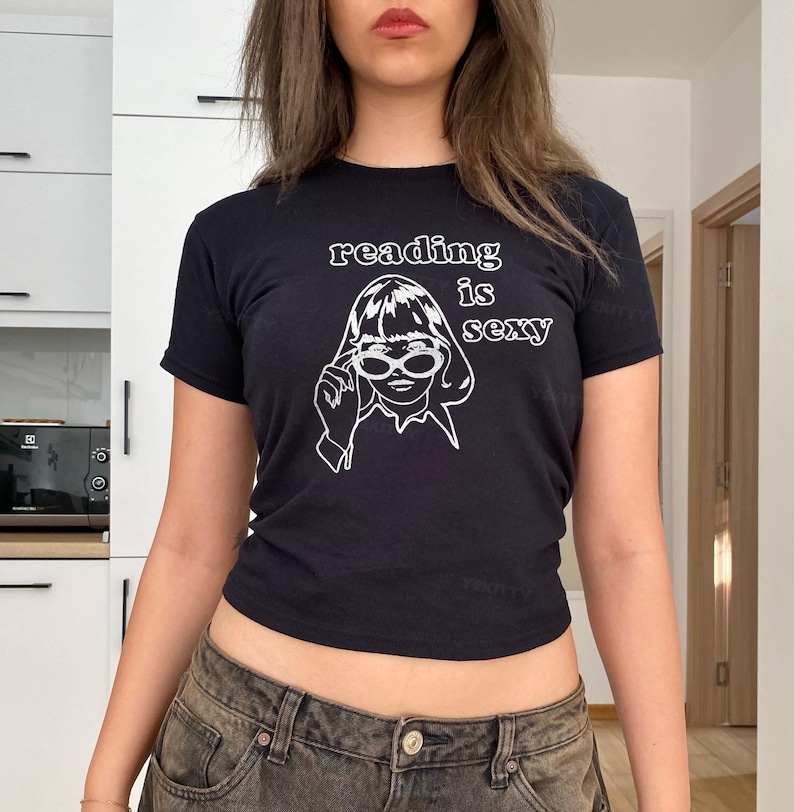 May include: Black t-shirt with the white text "reading is sexy" above a line drawing of a woman wearing sunglasses. The woman is touching her glasses. The shirt is short-sleeved.