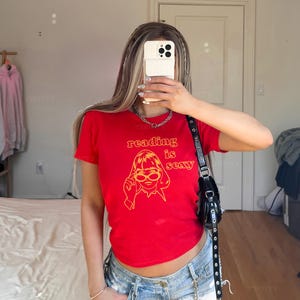 May include: Red t-shirt with the text "reading is sexy" in orange, along with a graphic of a woman wearing glasses. The shirt is paired with denim shorts. A black purse hangs from the shoulder.