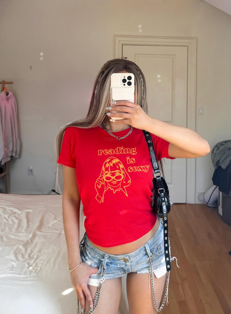 May include: Red t-shirt with the text "reading is sexy" in orange, along with a graphic of a woman wearing glasses. The shirt is paired with denim shorts. A black purse hangs from the shoulder.