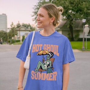 May include: A periwinkle blue short-sleeve t-shirt featuring a graphic print of a green-skinned ghoul in a white bikini lounging on a wooden beach chair under a yellow umbrella, with the orange text "HOT GHOUL SUMMER" printed above and below the graphic.