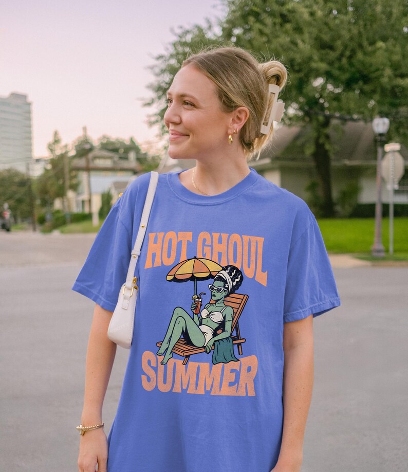 May include: A periwinkle blue short-sleeve t-shirt featuring a graphic print of a green-skinned ghoul in a white bikini lounging on a wooden beach chair under a yellow umbrella, with the orange text "HOT GHOUL SUMMER" printed above and below the graphic.