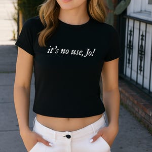 May include: Black crop top with the text "it's no use, Jo!" in white script. The shirt has short sleeves and a crew neck. The model is wearing white pants.