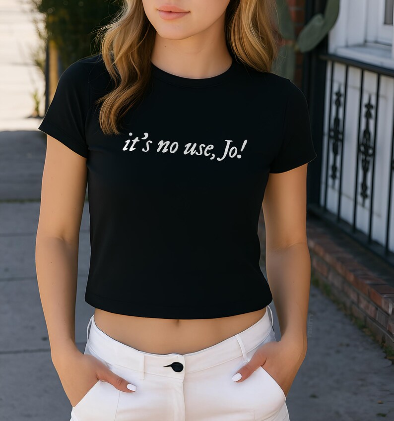 May include: Black crop top with the text "it's no use, Jo!" in white script. The shirt has short sleeves and a crew neck. The model is wearing white pants.