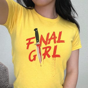 May include: Yellow t-shirt with the text "FINAL GIRL" in red with a bloody knife graphic.