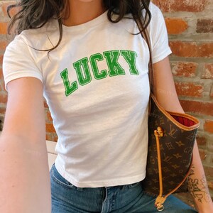 May include: White t-shirt with the word "LUCKY" in green block letters. The shirt is paired with blue jeans. A brown handbag with a gold zipper and a red interior is visible.
