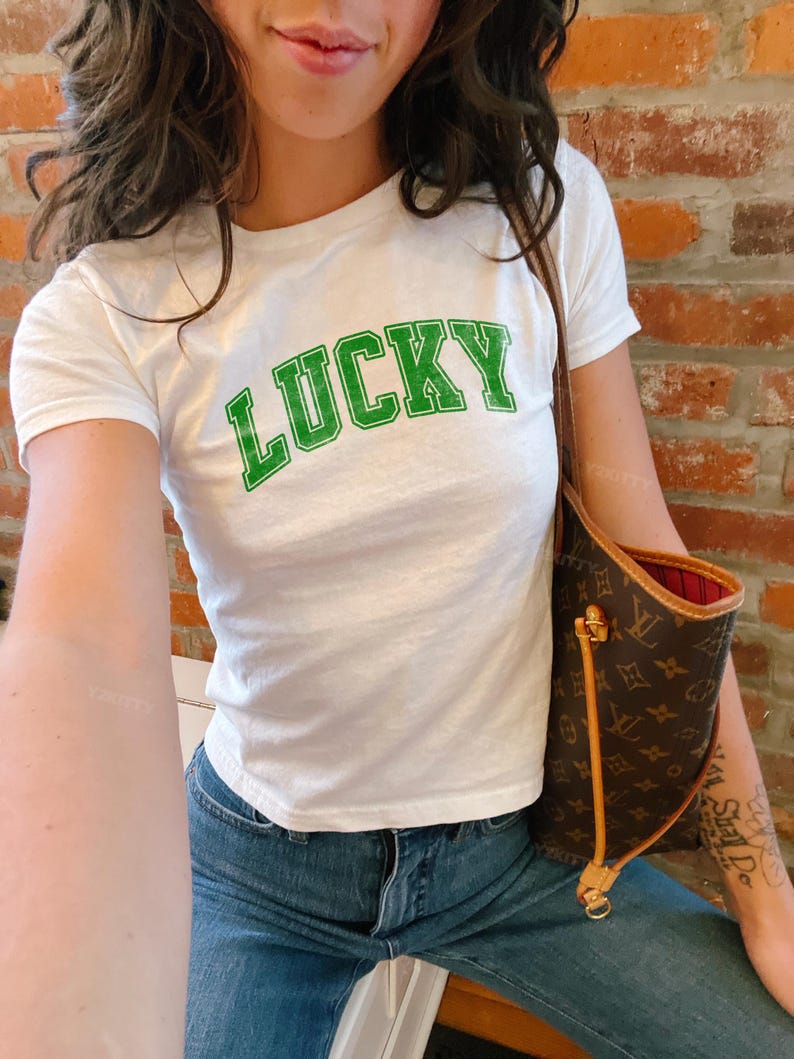 May include: White t-shirt with the word "LUCKY" in green block letters. The shirt is paired with blue jeans. A brown handbag with a gold zipper and a red interior is visible.
