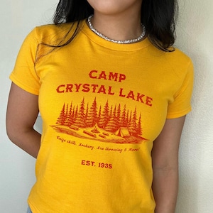 May include: Yellow t-shirt with a red graphic of a camp scene with trees, a tent, and a campfire. The text "CAMP CRYSTAL LAKE" is above the graphic, and the text "Knife skills. Archery. Axe throwing & More! EST. 1935" is below the graphic.