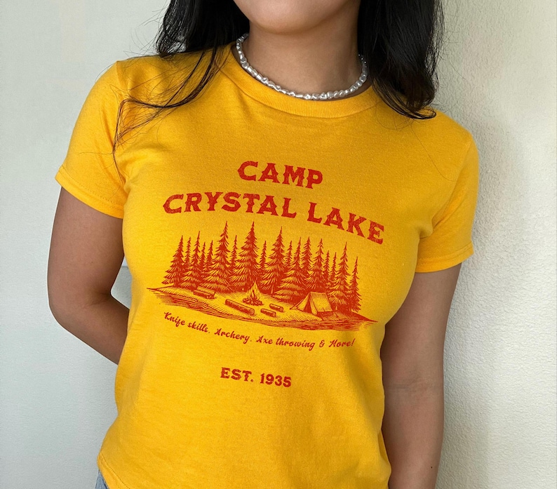 May include: Yellow t-shirt with a red graphic of a camp scene with trees, a tent, and a campfire. The text "CAMP CRYSTAL LAKE" is above the graphic, and the text "Knife skills. Archery. Axe throwing & More! EST. 1935" is below the graphic.