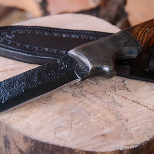 May include: A handcrafted hunting knife with a textured black blade and a wooden handle. The knife rests on a wooden surface, accompanied by a black leather sheath. The handle features a rich, brown wood grain.