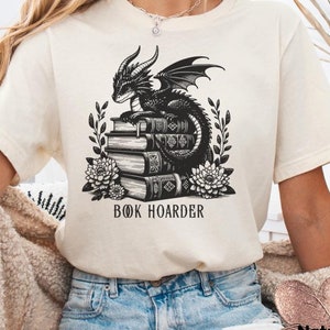 May include: A white t-shirt with a black and white illustration of a dragon sitting on a stack of books. The text "BOOK HOARDER" is printed below the illustration.