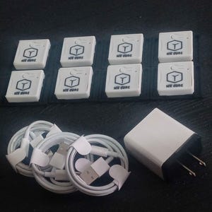 Puede incluir: Un conjunto de cables de carga blancos, ocho cajas blancas con el texto "ICE CURE" y un logotipo, y un adaptador de corriente blanco con clavijas negras. Los artículos están dispuestos sobre una superficie oscura.