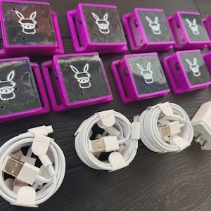 Puede incluir: Una colección de dispositivos electrónicos morados y negros con un logotipo de conejo, junto con cables de carga y adaptadores de corriente blancos. Los dispositivos tienen forma cuadrada con asa y pantalla.