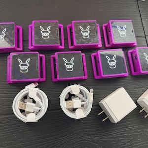 Puede incluir: Una colección de diez dispositivos cuadrados, morados y negros con un logotipo de conejo, junto con cables de carga y adaptadores de corriente blancos. Los dispositivos tienen forma rectangular con pantalla y marco morado.