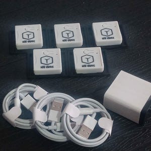 Może przedstawiać: Zestaw białych akcesoriów elektronicznych, w tym sześć małych kwadratowych pudełek z napisem "ICE CURE" i logo, kilka białych kabli USB oraz biały zasilacz z czarnymi bolcami. Ułożone na ciemnej powierzchni.