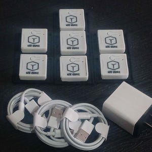 Puede incluir: Un conjunto de dispositivos electrónicos blancos, incluyendo pequeñas unidades cuadradas con la etiqueta "ICE CURE" y un logotipo, junto con cables de carga blancos y un adaptador de corriente. Los artículos están dispuestos sobre una superficie oscura.