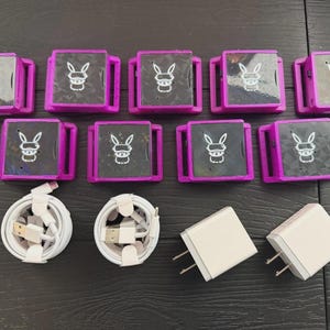 Puede incluir: Una colección de diez dispositivos electrónicos cuadrados, morados y negros, con un logotipo de conejo blanco, dispuestos sobre una superficie oscura. Se incluyen dos cables USB blancos y dos adaptadores de corriente blancos. Los dispositivos tienen un diseño con bisagras.
