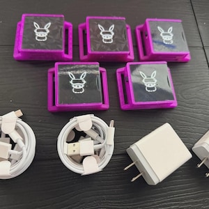 Puede incluir: Un surtido de accesorios electrónicos, que incluyen seis dispositivos cuadrados morados y negros con un logotipo de conejo, dos cables USB blancos y dos adaptadores de corriente blancos con clavijas. Los artículos están dispuestos sobre una superficie oscura.