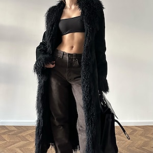 May include: A long black coat with a shaggy trim around the collar and cuffs. The coat is worn with brown trousers and a black top. A black handbag is also visible.