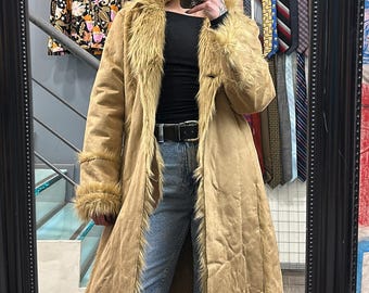 May include: A long, brown suede coat with a faux fur collar and trim. The coat has a long, flowing silhouette and is perfect for a cold winter day.