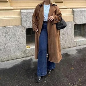 Puede incluir: Abrigo largo de ante marrón con diseño de doble botonadura. El abrigo se lleva sobre una camiseta blanca y unos vaqueros acampanados azul oscuro. Un bolso negro tejido y botines negros de punta cuadrada completan el conjunto.