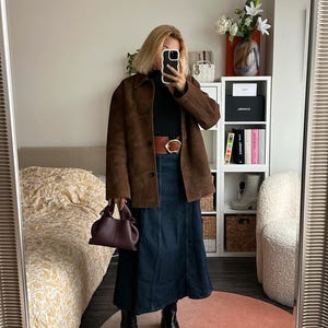 May include: A woman wearing a brown suede jacket, a black turtleneck sweater, a dark blue denim skirt, a brown belt with a gold buckle, and black heeled boots. She is holding a small burgundy handbag.