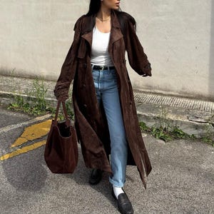 Könnte beinhalten: Eine Frau trägt einen braunen Wildledermantel mit einem weißen T-Shirt und einer blauen Jeans. Sie trägt eine braune Ledertasche.