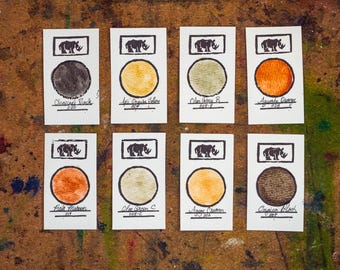 May include: Eight rectangular cards with ink swatches in various colors, including charcoal black, aqua orange, and beige cream. Each card features a rhino logo and color name. The cards are arranged on a wooden surface.
