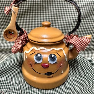 May include: A tan ceramic teapot shaped like a gingerbread person with a painted face, white icing trim, and a lid. It features a black metal handle adorned with red and white checkered fabric bows and a small wooden spoon with a painted face.