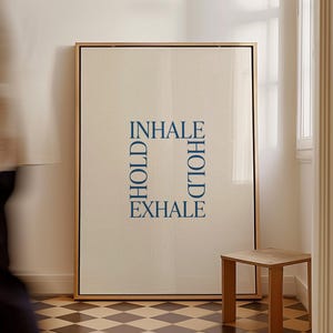Könnte beinhalten: Ein rechteckiges gerahmtes Wandbild mit hellbeigem Hintergrund und dunkelblauem Text in quadratischer Anordnung. Der Text lautet INHALE oben, EXHALE unten sowie HOLD an der linken und rechten vertikalen Seite des Quadrats.