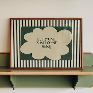 以下が含まれることがあります： 「EVERYONE IS WELCOME HERE」の文字が雲の形にデザインされたフレーム入りプリント。緑と白のストライプの背景に配置され、緑と木目調の棚に飾られています。