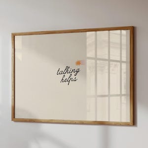Puede incluir: Un cuadro enmarcado con la frase "talking helps" en un estilo manuscrito, acompañado de una pequeña ilustración de un sol. El marco es de madera clara y el fondo es de color beige neutro.