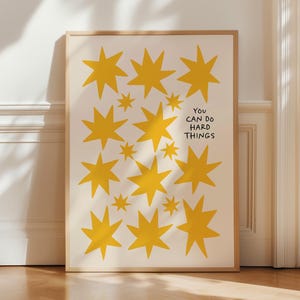Può includere: Una stampa artistica incorniciata con sfondo color crema e motivi a stella gialla. La stampa include il testo nero "YOU CAN DO HARD THINGS". L'opera è presentata in una cornice di legno, ideale per l'esposizione a parete.
