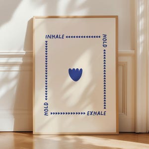 Könnte beinhalten: Gerahmter Kunstdruck mit minimalistischem Design. Das Kunstwerk zeigt die Wörter "INHALE", "HOLD" und "EXHALE" in Marineblau, angeordnet um eine zentrale blaue Blumenform. Der Hintergrund ist cremefarben.