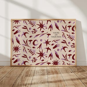 Può includere: Una stampa artistica incorniciata con sfondo beige e un motivo ripetuto di forme botaniche e celesti bordeaux e lavanda. Il testo "YOU'RE ON YOUR PATH" è in alto a destra. La cornice è in legno chiaro.