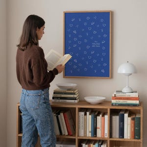 May include: A blue poster with white smiling face graphics and the text "ALL FEELINGS ARE OKAY" is displayed on a wall. A woman reads a book in front of a wooden bookshelf with books, a white lamp, and two white bowls.