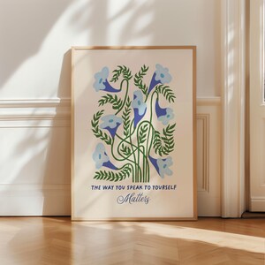 May include: A framed art print featuring a floral design with light blue flowers and green leaves. The artwork includes the text "THE WAY YOU SPEAK TO YOURSELF Matters" in blue and black font. The print is set against a cream background.