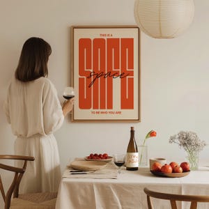 May include: A framed poster with the words "THIS IS A GOOD space TO BE WHO YOU ARE" in orange and black. A woman in a cream dress holds a glass of red wine. A table is set with fruit, plates, and a wine bottle.