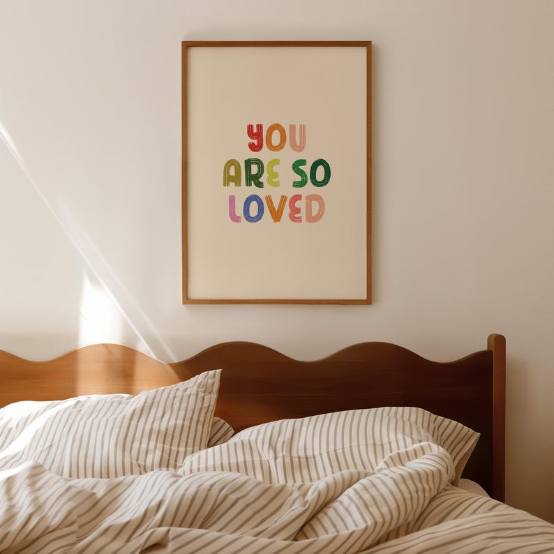 May include: A framed print with the text "YOU ARE SO LOVED" in colorful, stylized lettering. The print is hanging on a white wall above a bed with a brown headboard and striped bedding.